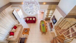 A spacious living area with a red sofa, a wooden coffee table, and a dining table with chairs. A unique chandelier hangs from the ceiling, and a spiral staircase is visible in the background. - Villa Lardos - Four Bedroom Villa, Sleeps 10 (Rhodes)