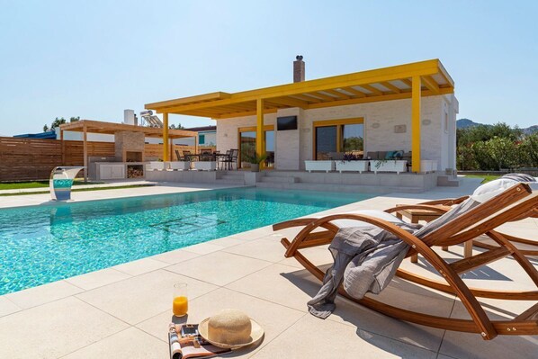 Un área junto a la piscina con un espacio de estar cubierto, sillas de descanso y zona de comedor.