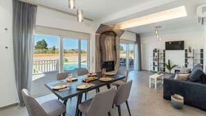 A spacious living room with a dining area, a fireplace, a TV, and a balcony with a pool. - Kolympia Petra - Two Bedroom Villa, Sleeps 6 (Kolympia)