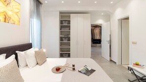 A modern hotel room with a neatly made bed, a wardrobe with shelves, a bedside table with a magazine, and a view into the hallway. - Kolympia Petra - Two Bedroom Villa, Sleeps 6 (Kolympia)