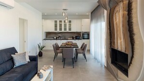 A modern living room with a sofa, dining table, and chairs. There is a kitchen in the background with white cabinets and a built-in oven. - Kolympia Petra - Two Bedroom Villa, Sleeps 6 (Kolympia)
