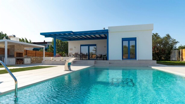 A modern pool area with a clear blue pool, a covered patio with outdoor furniture, and a white building with blue doors.