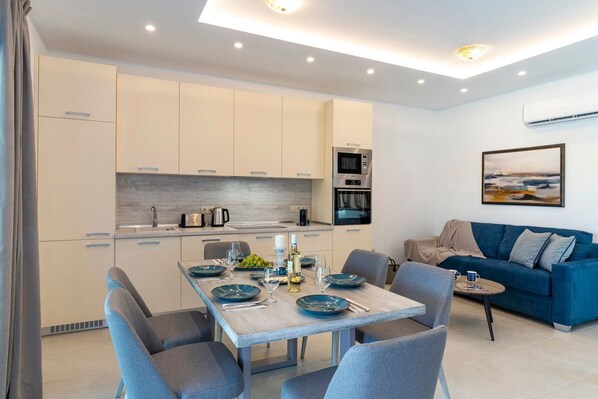 A modern kitchen with white cabinets, a dining table set with blue plates, and a blue sofa in the living area. - Kolympia Thalassa - Two Bedroom Villa, Sleeps 6 (Kolympia)