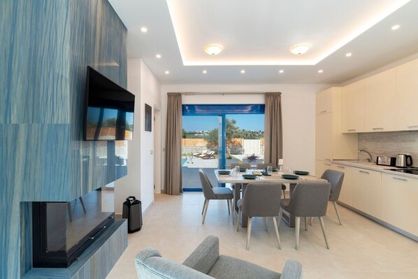 A modern living room with a flat-screen TV, a sofa, and a dining area with a table and chairs. - Kolympia Thalassa - Two Bedroom Villa, Sleeps 6 (Kolympia)
