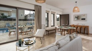 Ein geräumiger Wohnbereich mit einer großen Schiebeglastür, die zu einem Pool führt, ein Essbereich mit Holztisch und Stühlen sowie eine gemütliche Sitzecke mit einer Couch und einem kleinen runden Tisch.