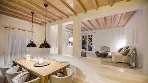 Un comedor con una mesa y sillas de madera, una lámpara colgante y vistas a una sala de estar con un sofá y chimenea.