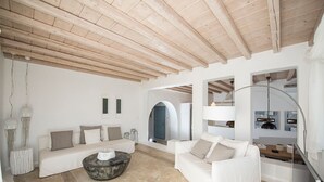 Amplia sala de estar con techo de madera alta, sofás blancos, una mesa de centro redonda y una cocina moderna visible a través de un arco.