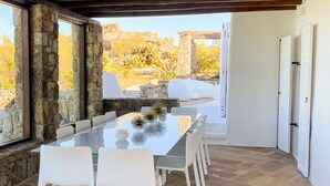 A spacious dining area with a glass table and white chairs, stone walls, and a view of a rocky landscape. - Villa Charoite - Six Bedroom Villa, Sleeps 12 (Thira)