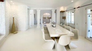 A modern, spacious dining area with a long white table and matching chairs, white walls, and a view into the kitchen. - Villa Charoite - Six Bedroom Villa, Sleeps 12 (Thira)