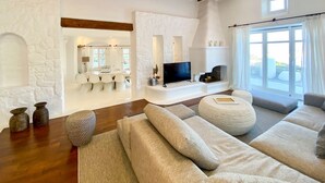A spacious living room with a stone fireplace, a flat-screen TV, and a large window with curtains. - Villa Charoite - Six Bedroom Villa, Sleeps 12 (Thira)