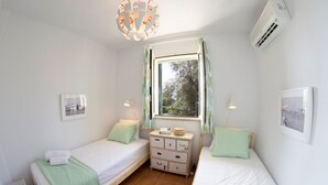 A small, well-lit bedroom with two single beds, a nightstand, a window with curtains, and an air conditioning unit. - Villa Arithmos - Three Bedroom Villa, Sleeps 6 (Lefkada)
