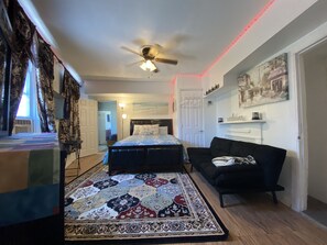 Un dormitorio con una cama, ventilador de techo, alfombra estampada, sofá negro y chimenea.