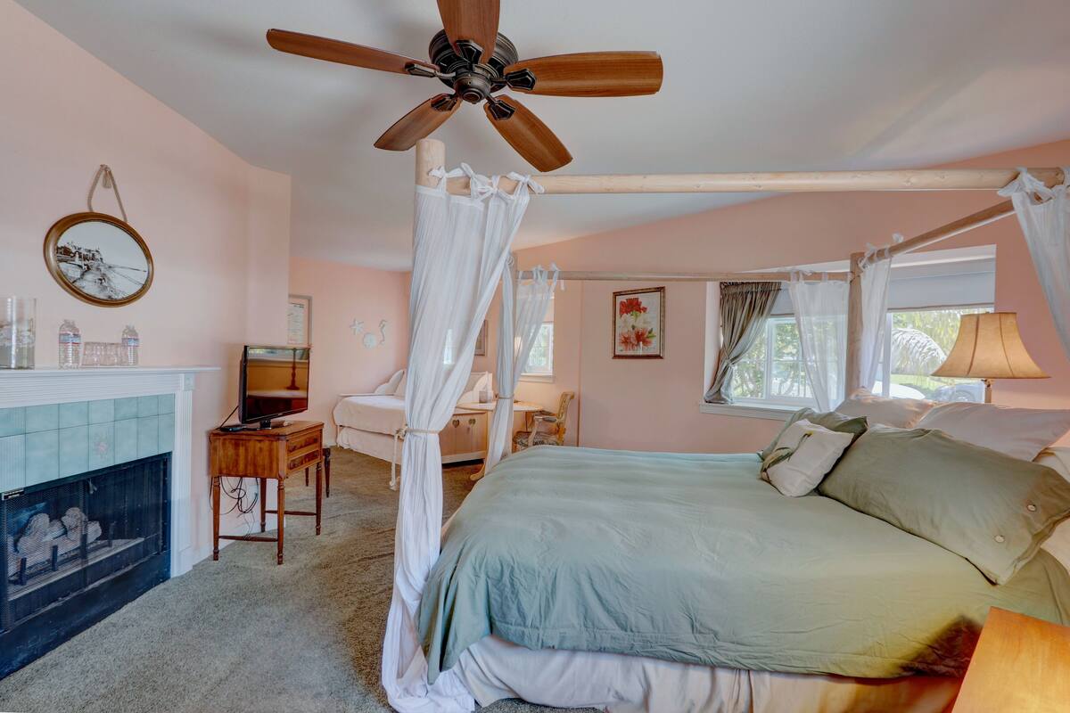 A bedroom with a four-poster bed, a ceiling fan, a fireplace, and a television.
