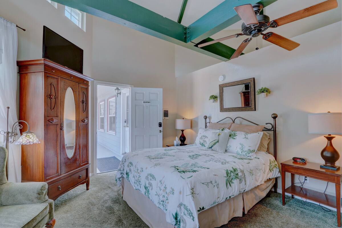 A bedroom with a bed, a wardrobe, a ceiling fan, a bedside table with a lamp, and a door leading to another room.
