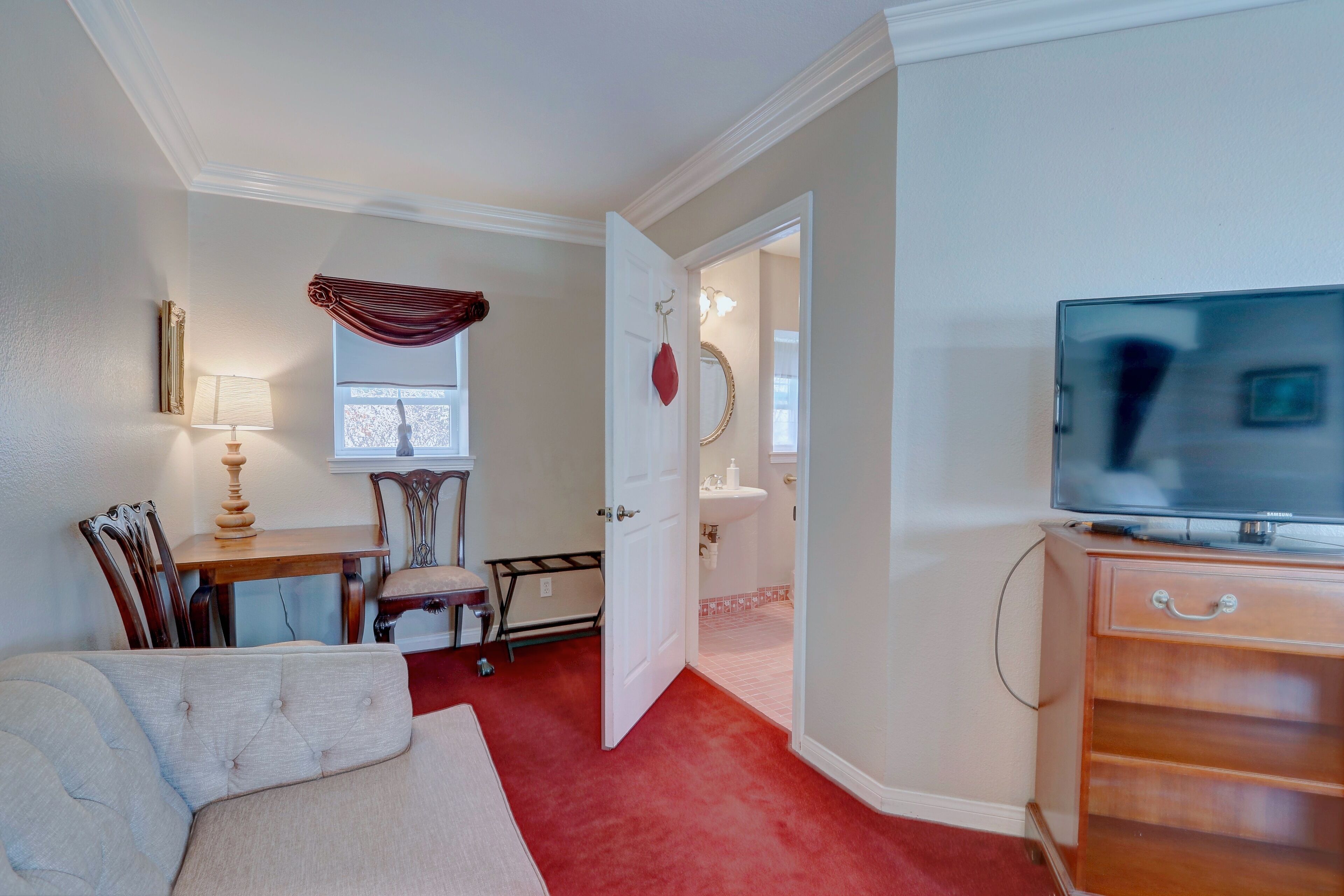 A room with a sofa, a television on a wooden cabinet, a dining table with chairs, and a door leading to a bathroom.