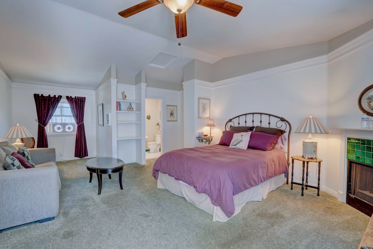 A bedroom with a bed, a ceiling fan, a fireplace, and a small table.