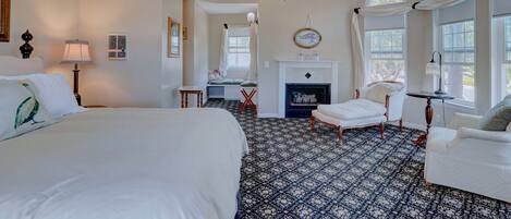 A spacious bedroom with a large bed, a desk, a fireplace, and a seating area with a chair and a sofa.