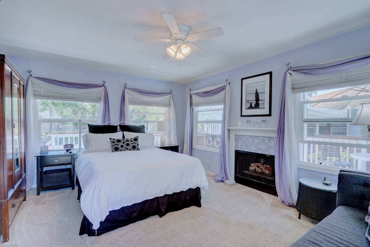 A bedroom with a large bed, a ceiling fan, a fireplace, and a view of the outside.