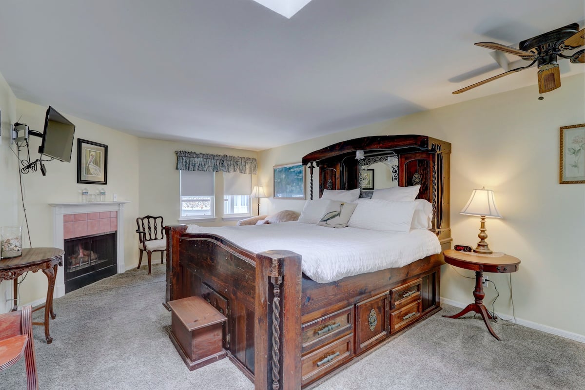 A bedroom with a large bed, a fireplace, a ceiling fan, and a small table with a lamp.