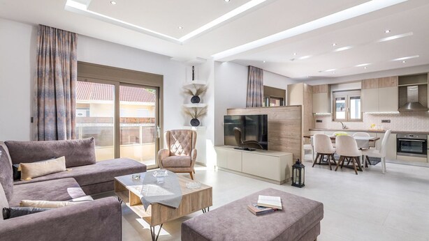 Un salon moderne comprenant un canapé, un fauteuil et un pouf, un coin repas avec des chaises, une cuisine avec un téléviseur et une grande fenêtre avec des rideaux.
