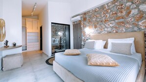 A modern bedroom with a large bed, a stone accent wall, a built-in wardrobe, and a small seating area. - Villa Arnassica - Three Bedroom Villa, Sleeps 6 (Ialysos)