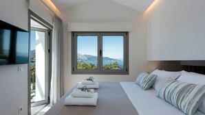 A modern bedroom with a large bed, a TV mounted on the wall, and a window offering a scenic view. - Villa Chloris - Three Bedroom Villa, Sleeps 8 (Kontokali)