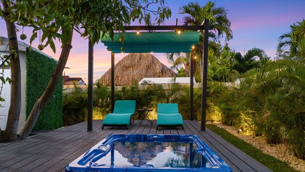 Un bain  à remous sur une terrasse en bois, avec deux chaises longues sous une pergola, entouré de verdure et avec une montagne en arrière-plan.