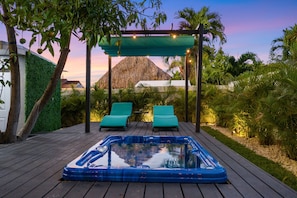 A hot tub on a wooden deck with two lounge chairs under a pergola, surrounded by greenery and a mountain in the background.