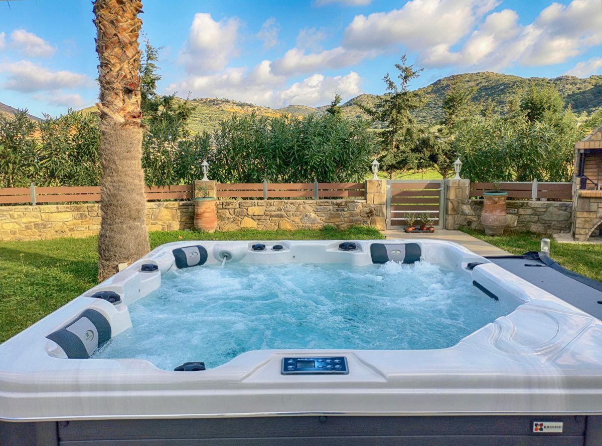 A hot tub with a control panel, surrounded by a stone wall, a wooden fence, and lush greenery.