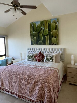 A bedroom with a tufted headboard, a patterned bedspread, and a cactus artwork on the wall.