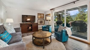A living room with a television, sofa, chairs, a coffee table, and a sliding glass door leading to a deck. - Whitefish Lake Condo, 14BD, hillside! (White)