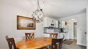 A dining area with a wooden table and chairs, a chandelier, a framed picture on the wall, and a kitchen in the background. - Whitefish Lake Condo, 14BD, hillside! (White)