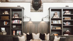 Una habitación con techo de madera, dos estanterías con libros y adornos, una cabeza de animal montada y una zona de estar con cojines.