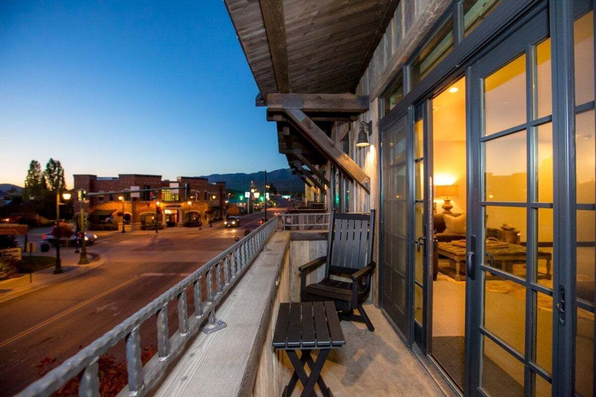 Un balcón con vistas a una calle y montañas, muebles de madera y un ventanal que permite ver el interior.