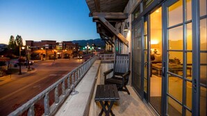 Un balcón con vistas a una calle y montañas, muebles de madera y un ventanal que permite ver el interior.