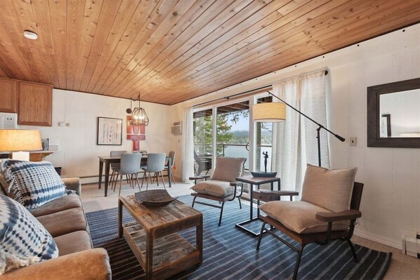 A cozy living room with a wooden ceiling, a dining area with chairs and a table, and a large window with curtains. - Waterfront, Whitefish Lake Condo, 2CD (Whitefish)