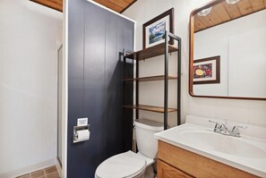A bathroom with a white toilet, a white sink, and a wooden vanity. There is a shelf unit above the toilet and a framed picture on the wall. - Waterfront, Whitefish Lake Condo, 2CD (Whitefish)