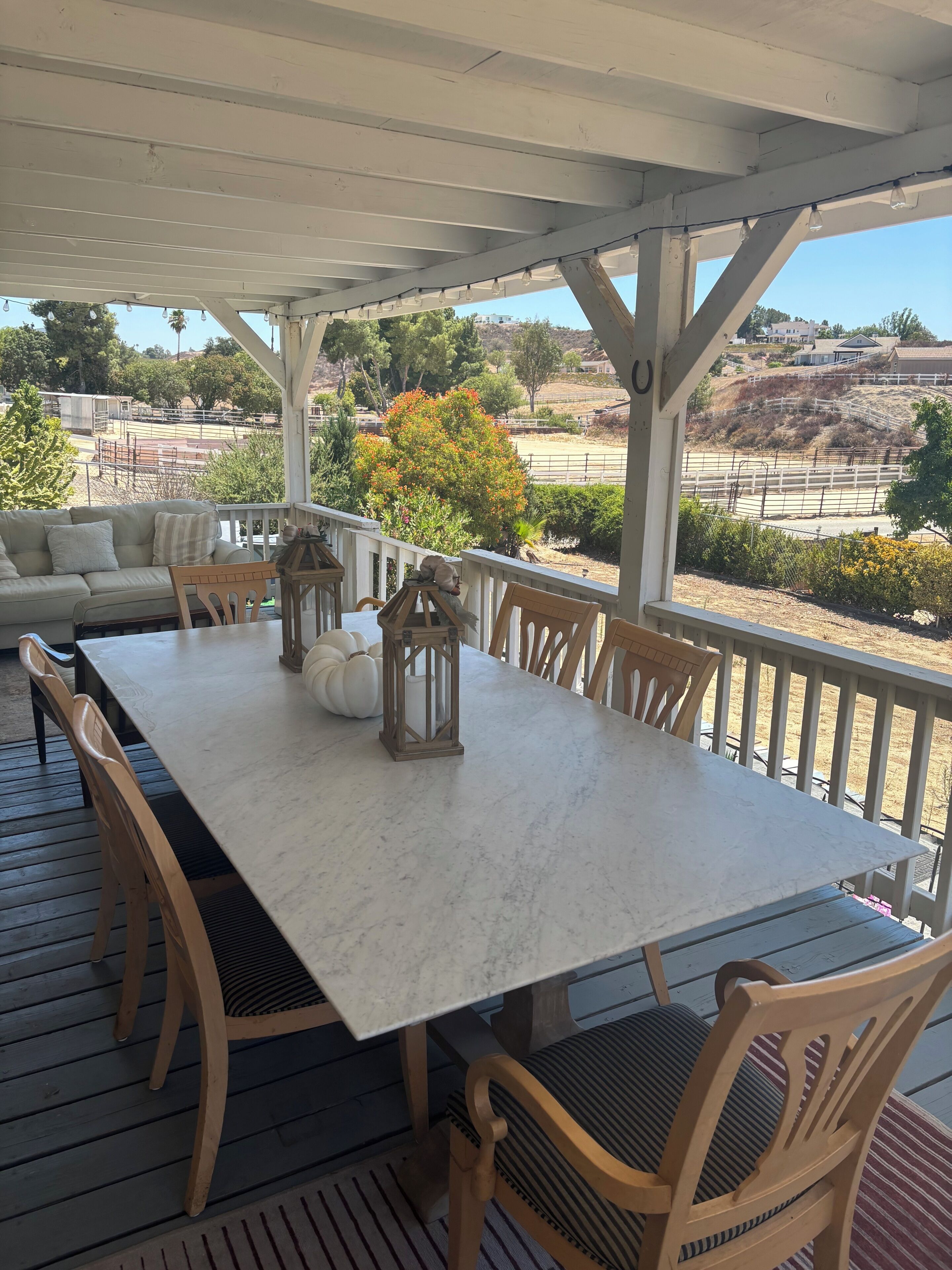 Un espace extérieur abrité, aménagé pour les repas, avec une table blanche et des chaises en bois, donnant sur une propriété clôturée, agrémentée d’arbres et de collines.