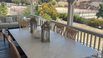 Un espace extérieur abrité, aménagé pour les repas, avec une table blanche et des chaises en bois, donnant sur une propriété clôturée, agrémentée d’arbres et de collines.