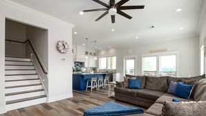 A spacious living area with a gray sofa, a blue ottoman, and a ceiling fan. A kitchen with white cabinets and a blue island is visible in the background. - Brand NEW Beach House Coastal Point  (Bolivar Peninsula)