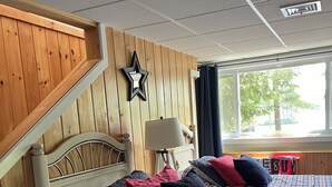 A room with two beds, wooden walls, a star decoration, a lamp, and a window with a view of trees.