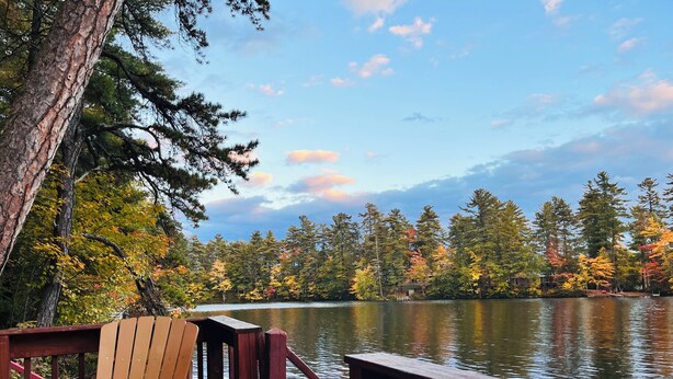 A wooden deck with chairs and a table, overlooking a calm lake surrounded by trees with autumn foliage.
