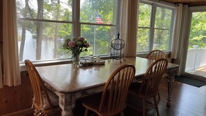 A dining area with a wooden table and chairs, large windows with curtains, and a vase of flowers.