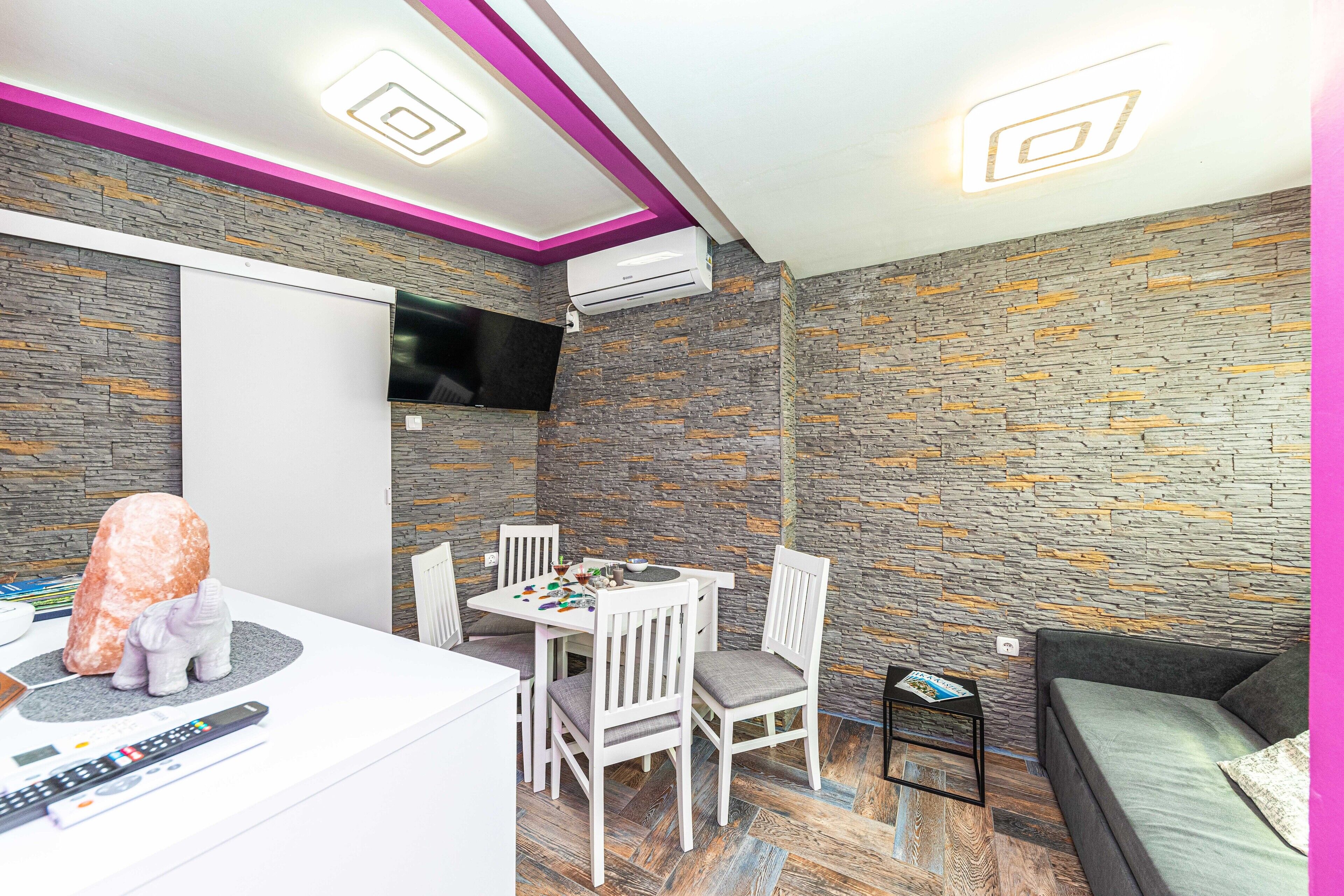 Une chambre moderne avec un mur décoré d’un motif imitant la pierre, un téléviseur à écran plat fixé au-dessus d’une table à manger avec des chaises, et un canapé agrémenté d’un coussin.