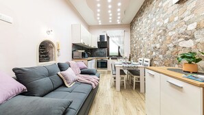 A modern living room with a grey sofa, a dining area with a wooden table, and a kitchen with stone wall backsplash.