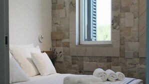 Een net opgemaakt bed met witte lakens en opgerolde handdoeken, een stenen muur en een raam met zonwering.
