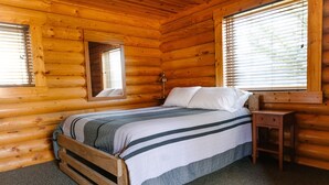 A wooden cabin bedroom with a bed, a nightstand, a lamp, and a window with blinds.