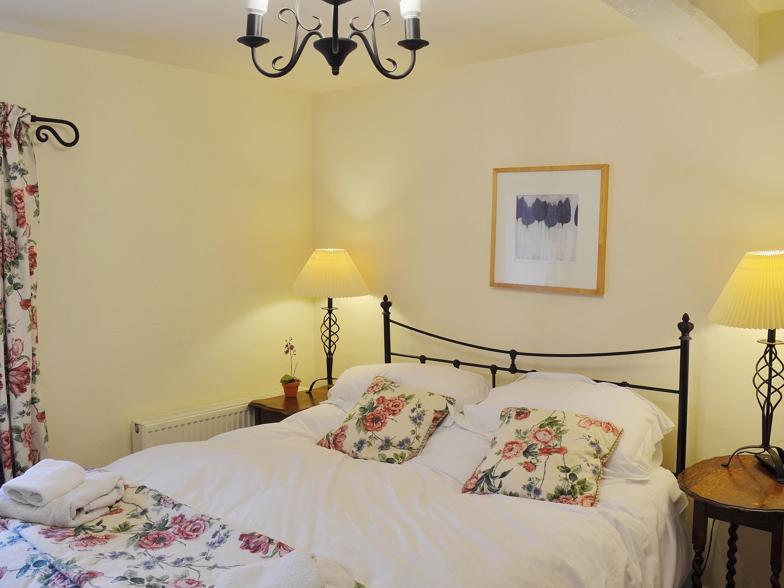 A neatly made bed with floral bedding, a chandelier, and a framed picture on the wall.