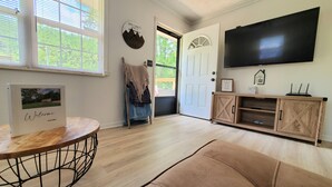 A room with a wooden floor, a round table with a welcome sign, a couch, a TV mounted on the wall, and a door leading outside. - Private/Between PF & Knox/DOG friendly/creek/large yard (Seymour)