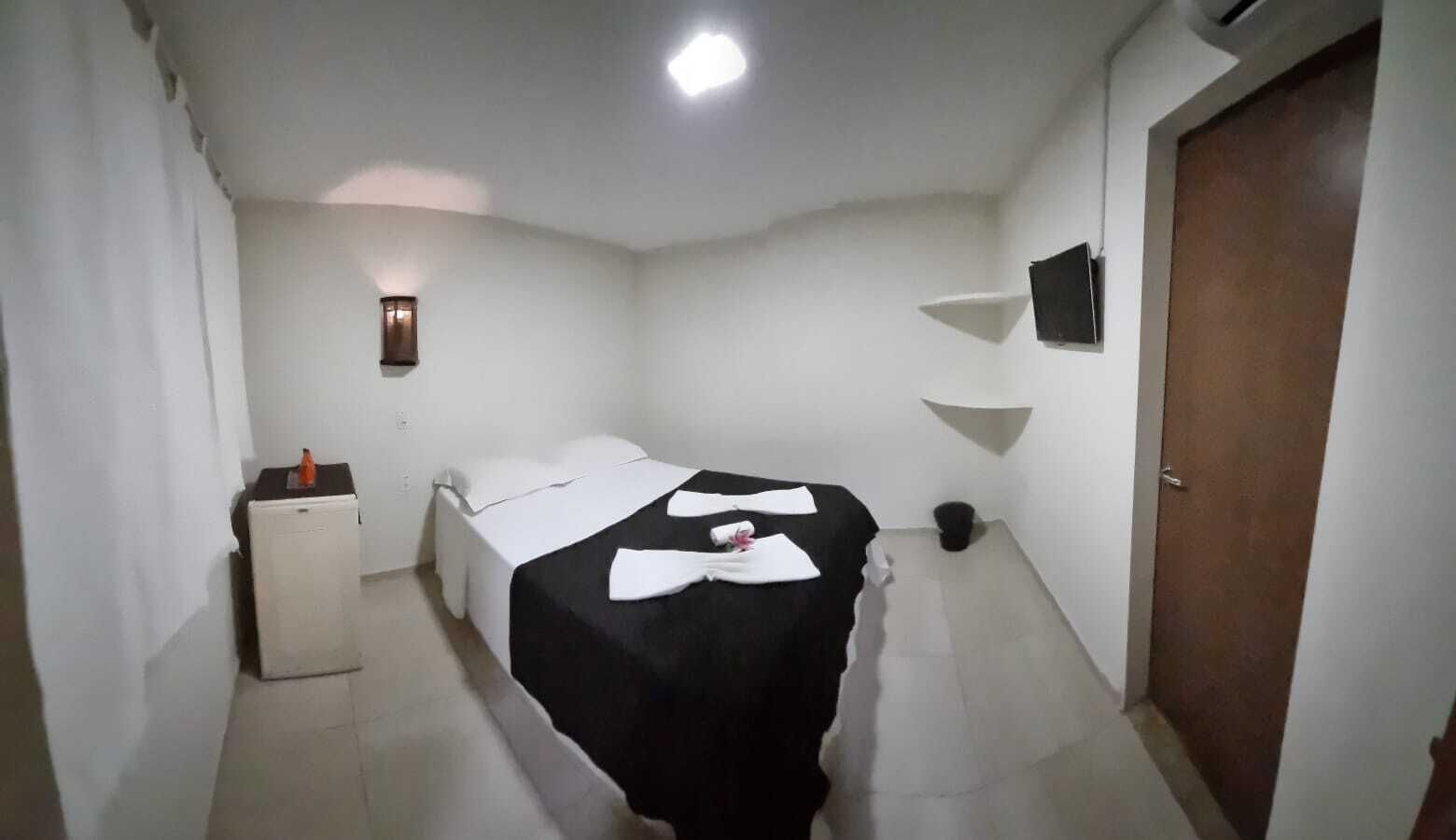 Quarto pequeno e limpo com uma cama, uma mesinha e um abajur, além de uma porta.